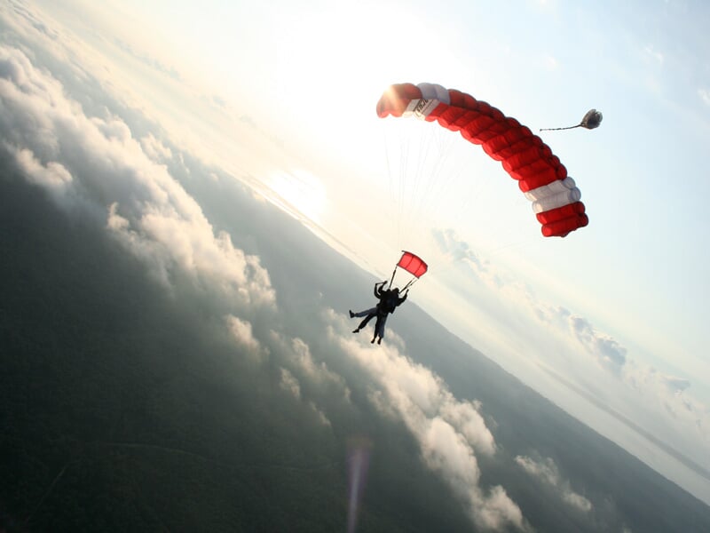 Parachutiste en tandem avec parachute rouge et blanc, descendant au-dessus des nuages au coucher du soleil.