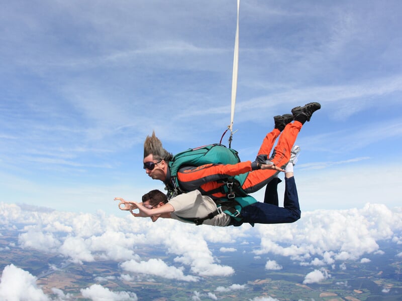 Deux personnes en chute libre en tandem, avec harnais, sur fond de ciel bleu et nuages blancs.