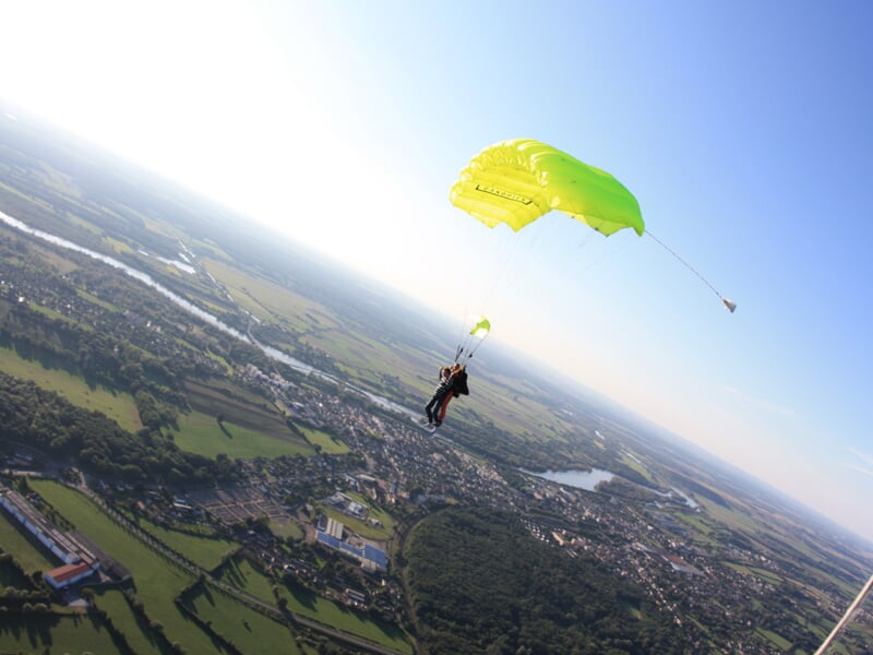Parachutiste en tandem avec parachute vert clair, survolant une campagne avec rivières et villages.