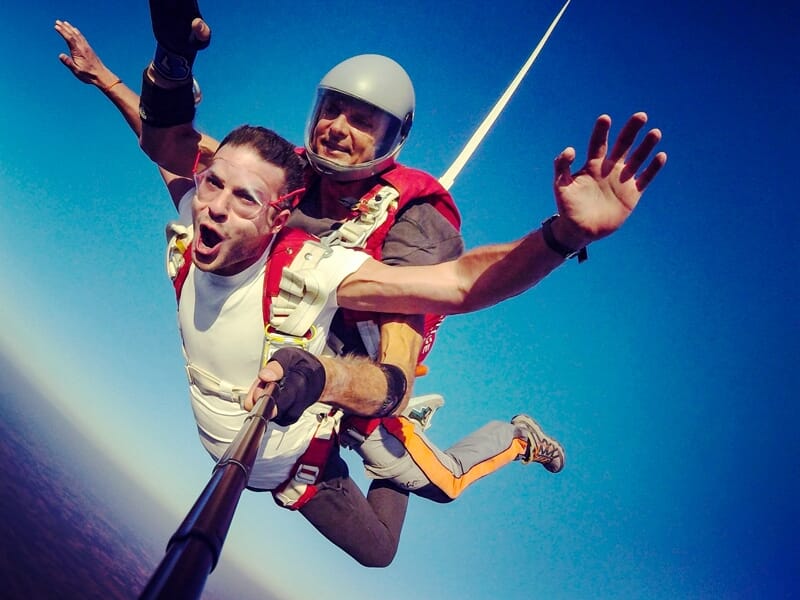 Deux hommes en chute libre en tandem, souriants, avec un ciel bleu clair en arrière-plan.