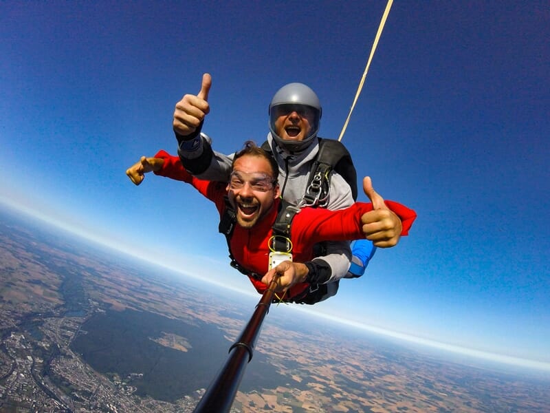 Deux hommes en chute libre en tandem, souriants, faisant un pouce levé avec un paysage vu du ciel.