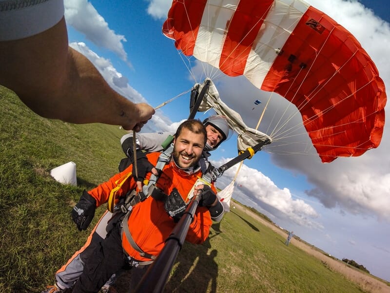 Parachutistes en tandem au sol, souriants, avec un parachute rouge et blanc déployé au-dessus d'eux.