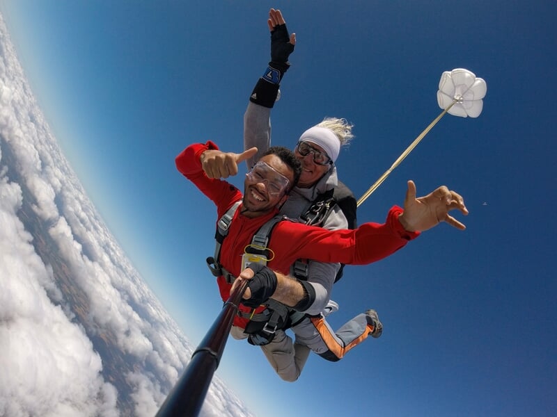 Deux hommes en chute libre en tandem, souriants, avec un petit parachute de secours blanc déployé derrière eux.