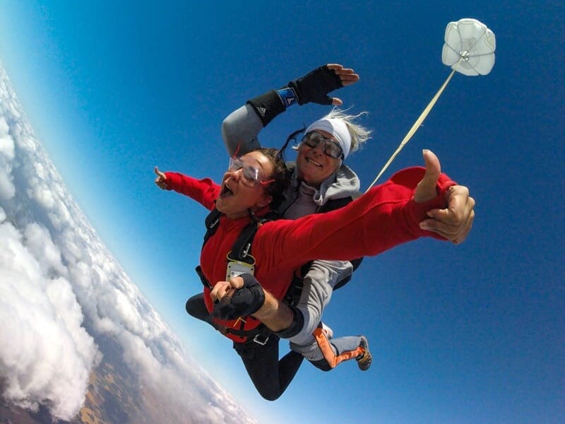 Deux hommes en chute libre en tandem, souriants, avec un petit parachute de secours blanc déployé en arrière-plan.