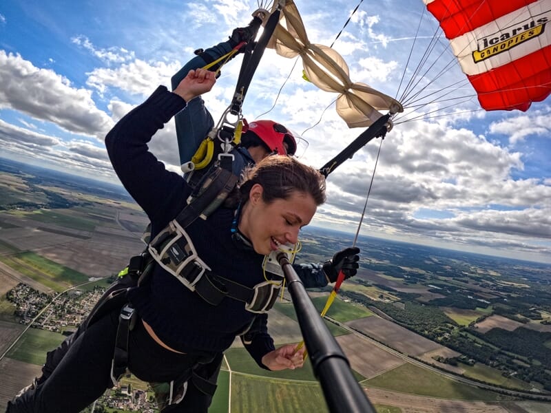 Femme en chute libre en tandem, souriante, tenant une perche selfie avec un parachute rouge et blanc déployé.