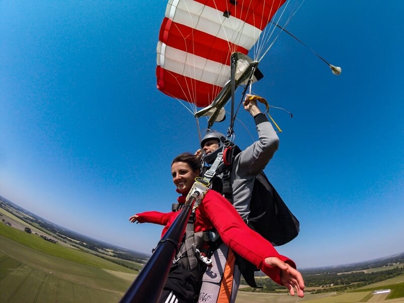 Femme en chute libre en tandem, souriante, tenant une perche selfie avec un parachute rouge et blanc déployé.