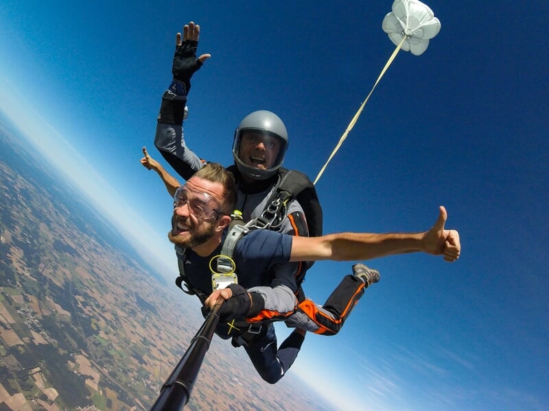Deux hommes en chute libre en tandem, souriants, avec un petit parachute de secours blanc déployé derrière eux.