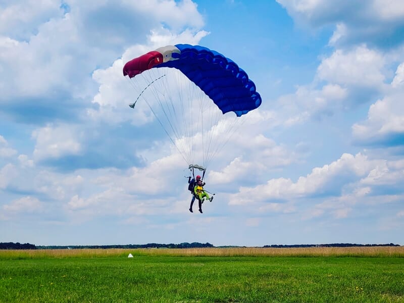 Parachutiste atterrissant sur un champ vert avec un parachute bleu, blanc et rouge déployé au-dessus.