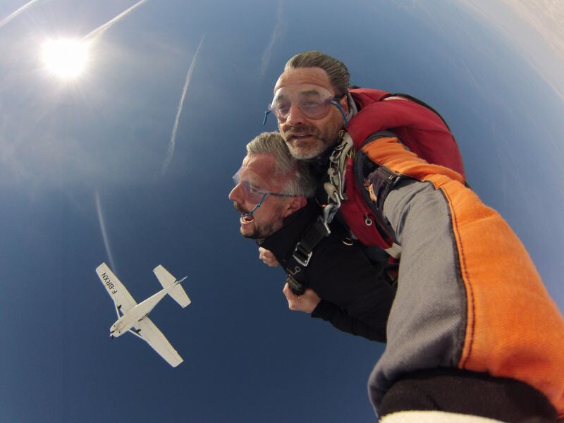 Deux hommes en tandem effectuent un saut en parachute sous un ciel bleu avec un avion en arrière-plan.