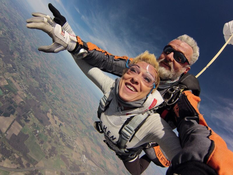 Deux personnes en parachute en tandem survolent un paysage rural avec ciel bleu et sourires de joie et d'excitation.