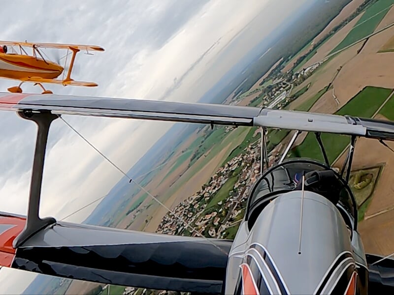 Vue en vol de deux avions biplans au-dessus d'un paysage rural avec champs et village sous un ciel nuageux.