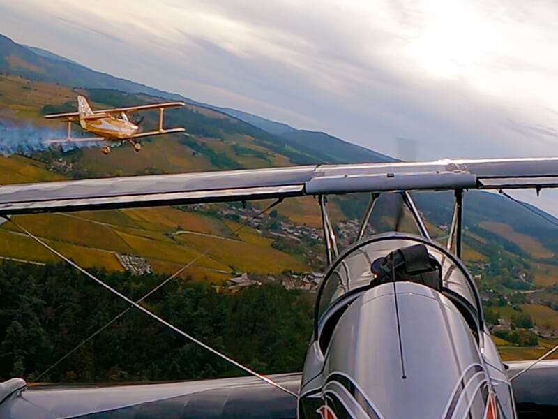 Deux avions biplans en vol près d'une colline boisée avec un village et des vignes en arrière-plan sous un ciel couvert.