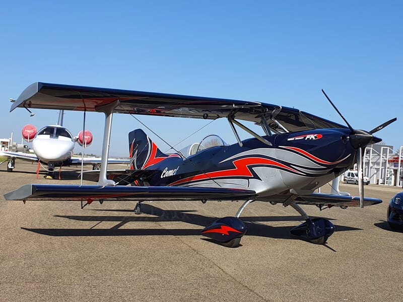 Avion biplan noir et rouge garé sur une piste d'aéroport sous un ciel bleu clair avec un autre avion en arrière-plan.