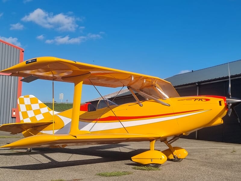 Petit avion jaune vif avec des motifs blancs garé sur une piste près d'un hangar sous un ciel bleu avec quelques nuages.