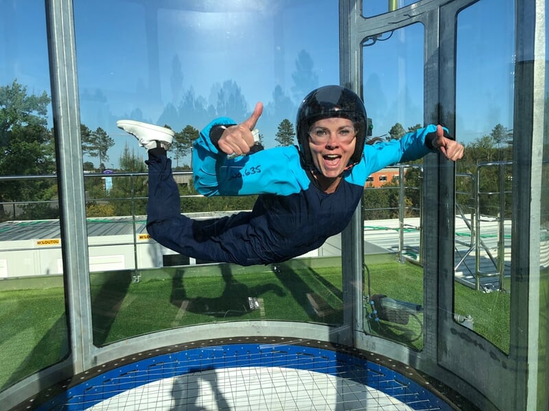 Femme en chute libre simulée : Une femme en combinaison de parachutisme bleue et noire sourit et montre un pouce en l'air dans une soufflerie. Elle porte un casque de protection et des lunettes. Le pa