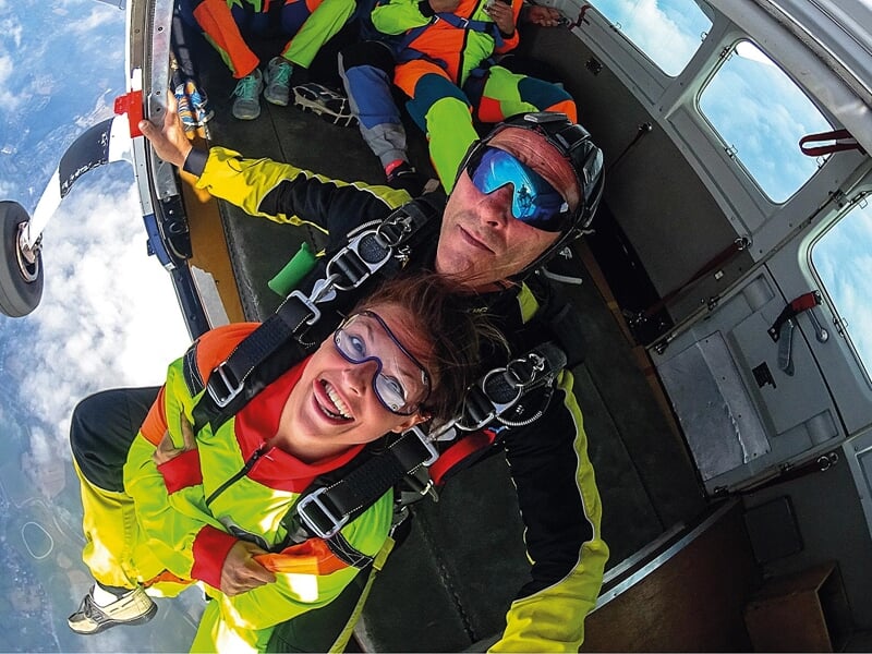 Préparation pour un saut en tandem en avion : Un duo de parachutistes en combinaison multicolore se prépare à sauter depuis un avion. La femme affiche un sourire large et excité tandis que l'instructe