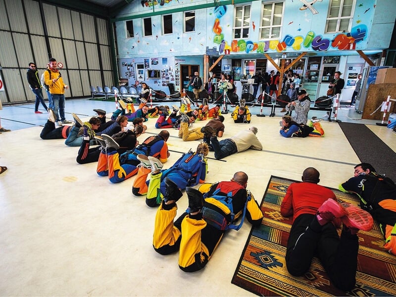 Exercice au sol pour parachutistes : Un groupe de parachutistes en combinaison est allongé au sol, pratiquant les positions de chute libre dans un hangar. L'instructeur donne des conseils, tandis que 