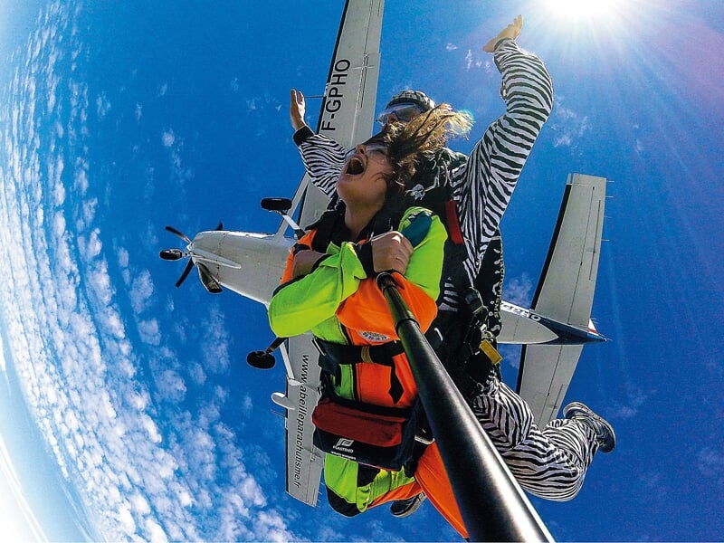 Chute libre en tandem au-dessus de l'avion : Une femme et son instructeur en combinaison colorée sautent en tandem d'un avion, tenant une perche à selfie. Le ciel est bleu avec quelques nuages épars, 