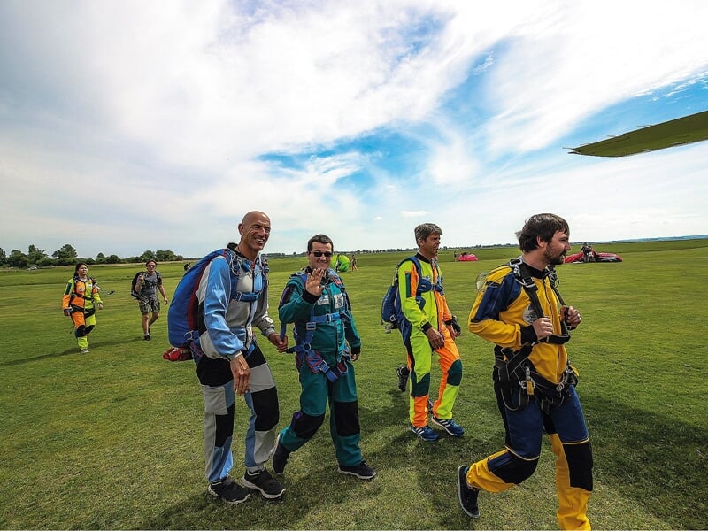 Retour sur terre après un saut en parachute : Un groupe de parachutistes en combinaison multicolore marche sur une pelouse après avoir atterri. Certains sourient et saluent la caméra, heureux et entho