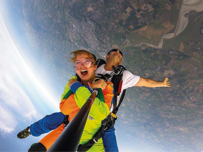 Chute libre en tandem au-dessus de la campagne : Une femme et son instructeur en combinaison de parachutisme colorée chutent librement au-dessus d'un paysage de campagne. Elle tient une perche à selfi