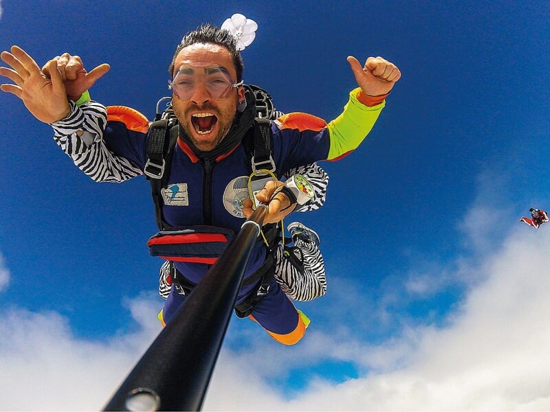 Parachutiste en chute libre solo : Un homme en combinaison de parachutisme colorée chute librement, tenant une perche à selfie. Il montre des signes de main et sourit largement, avec le ciel bleu et d