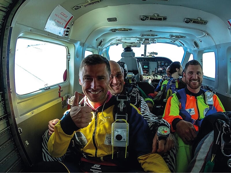 Intérieur d'un avion de parachutisme : Un groupe de parachutistes assis à l'intérieur d'un avion, prêt à sauter. L'un des parachutistes, en combinaison jaune, sourit et montre un pouce en l'air. L'amb