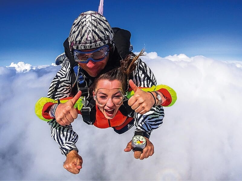 Parachutisme en tandem au-dessus des nuages : Une femme et son instructeur en combinaison colorée chutent librement au-dessus des nuages. Tous deux sourient et montrent des pouces en l'air, exprimant 