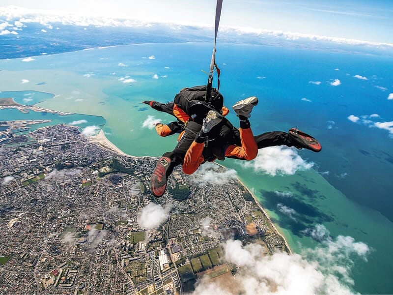 Chute libre en tandem au-dessus d'une ville côtière : Deux parachutistes en tandem, vêtus de combinaisons noires et oranges, chutent librement au-dessus d'une ville côtière. La vue offre un panorama s