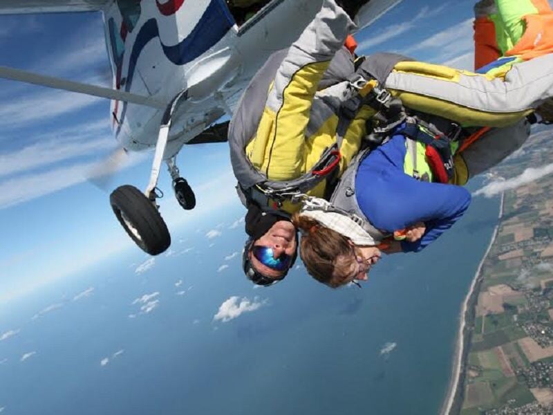 Sortie d'avion pour un saut en parachute : Un duo de parachutistes en tandem vient de quitter un avion en vol, prêt à commencer leur chute libre. La mer et le littoral sont visibles en arrière-plan so