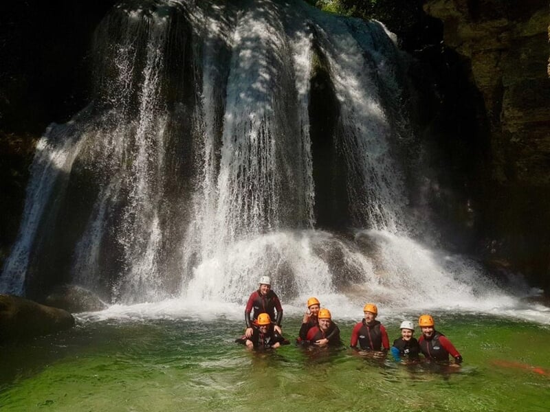 Groupe de personnes en tenue de canyoning posant dans une eau claire au pied d'une cascade impressionnante.
