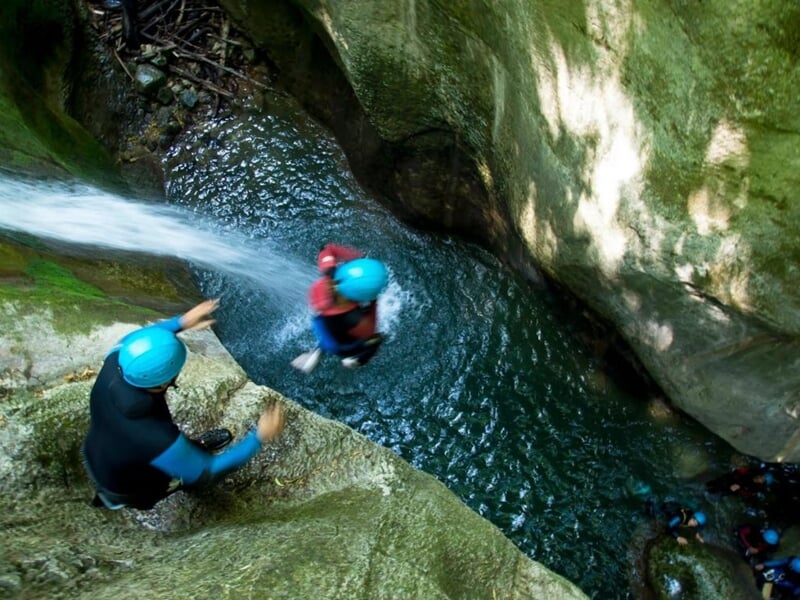 Personne en casque bleu sautant dans un bassin d'eau entouré de rochers, accompagnée d'un guide.
