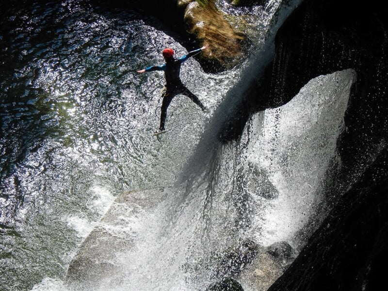 Personne en combinaison noire sautant dans une rivière depuis une falaise, sous une cascade d'eau.