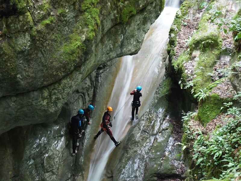 Trois personnes en casque bleu et combinaison noire progressant le long d'une cascade rocheuse en canyoning.