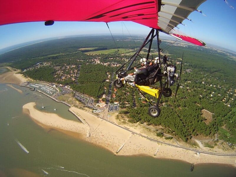 ULM volant au-dessus d'une marina et d'une plage animée, avec une vue sur une ville côtière entourée de forêt et de mer