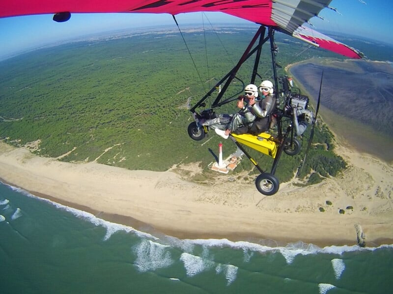 Vol en ULM au-dessus d'une plage bordée de forêt, avec un phare rouge et blanc visible en dessous. Scène de littoral et d'aviation légère