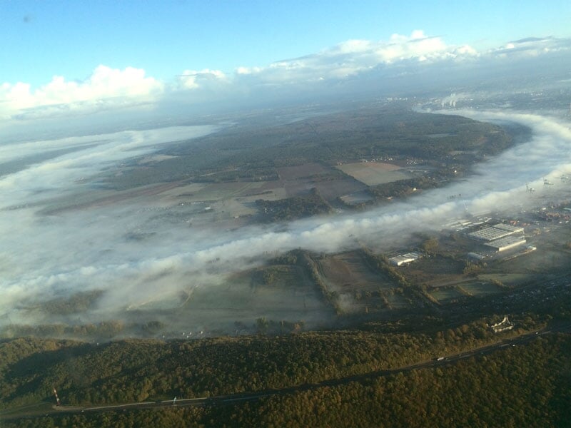 Vue aérienne d'un paysage avec des nuages bas sur des champs, des forêts et des bâtiments, depuis le cockpit d'un avion.