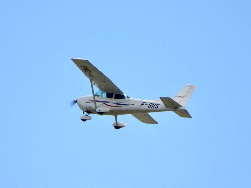 Petit avion blanc à ailes hautes en vol, avec un ciel bleu dégagé en arrière-plan.