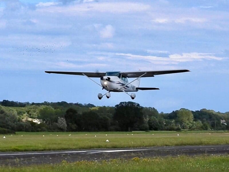 Un petit avion blanc à ailes hautes décollant d'une piste avec des arbres et un ciel bleu clair en arrière-plan.