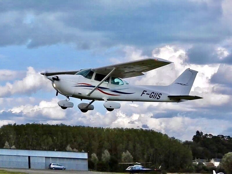Petit avion blanc avec des rayures bleues et rouges volant bas au-dessus d'un terrain avec des bâtiments et des arbres en arrière-plan.