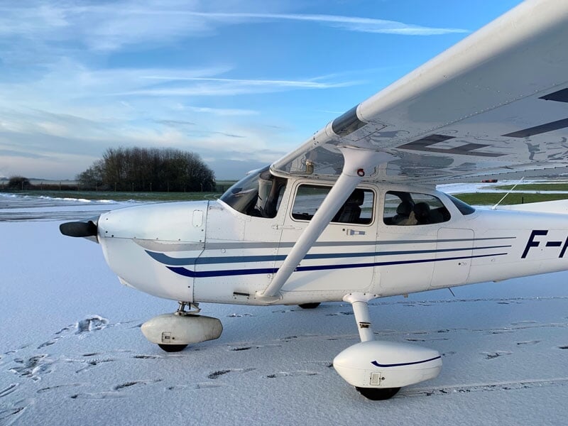 Petit avion blanc stationné sur une piste enneigée, avec des traces dans la neige et des arbres dénudés en arrière-plan.