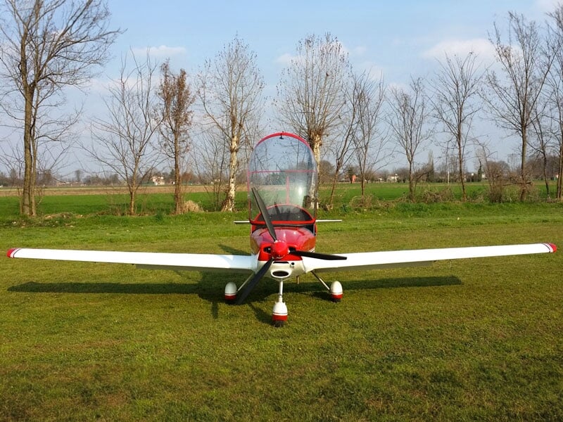 un avion ULM rouge au cockpit ouvert posé dans un champ d'herbe verte