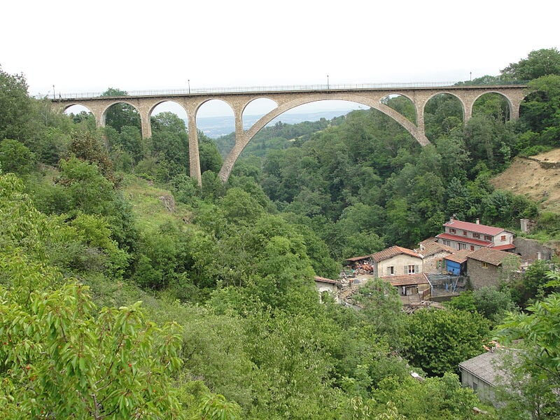Un pont en arc en béton surplombant une vallée verdoyante avec des maisons au pied de la colline.