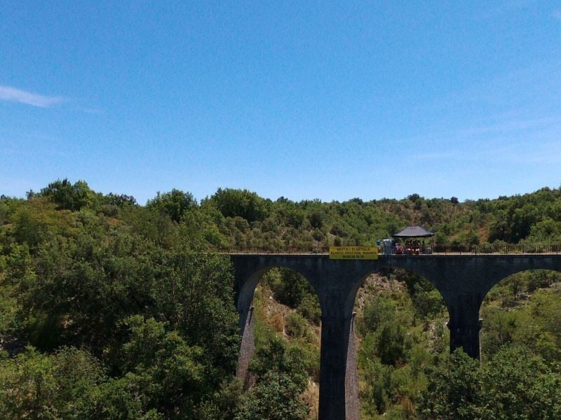 Viaduc en pierre dans un paysage forestier, avec une plateforme de saut à l'élastique équipée d'un auvent noir et d'une bannière jaune, sous un ciel bleu dégagé