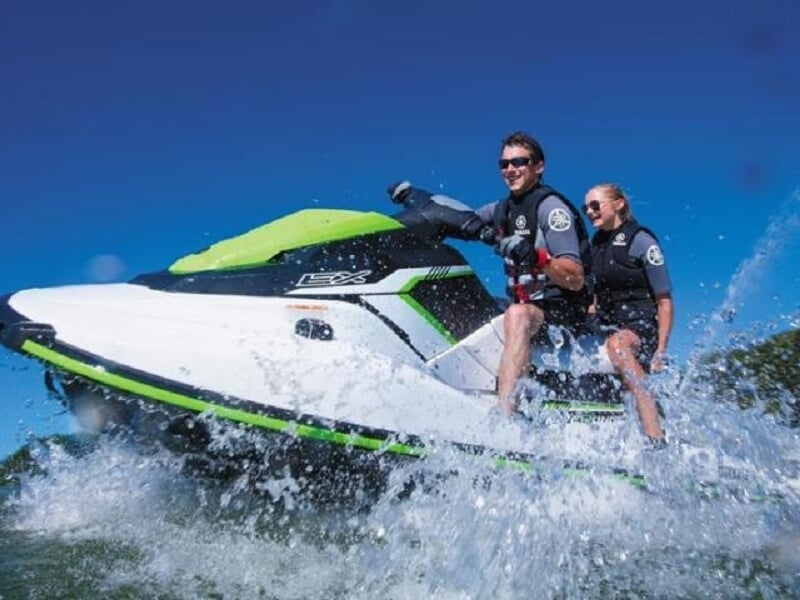 Deux personnes souriantes sur un jetski blanc et vert qui avance rapidement sur l'eau avec éclaboussures.