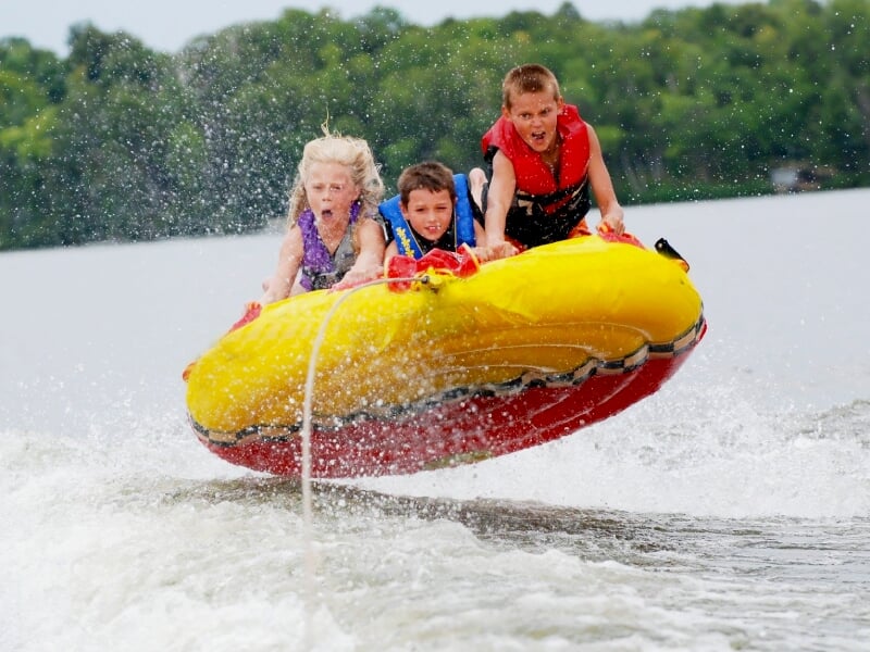 Trois enfants sur une bouée jaune tractée sur l'eau, éclaboussures autour, ambiance joyeuse et sportive.
