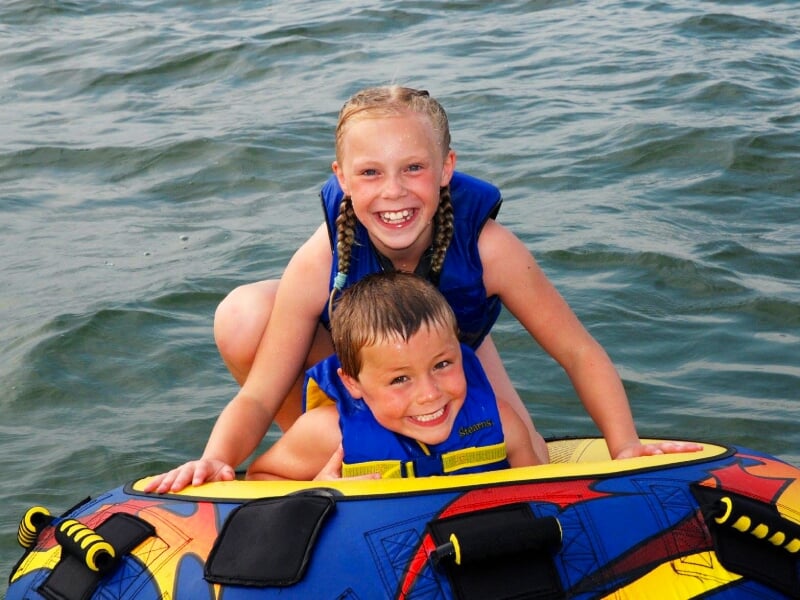Deux enfants souriants en gilets de sauvetage bleus sur une bouée colorée flottant sur l'eau calme.