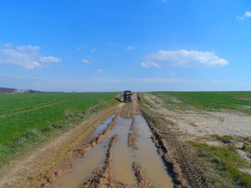 Chemin boueux en campagne avec un véhicule tout-terrain au loin sous un ciel bleu clair et quelques nuages.