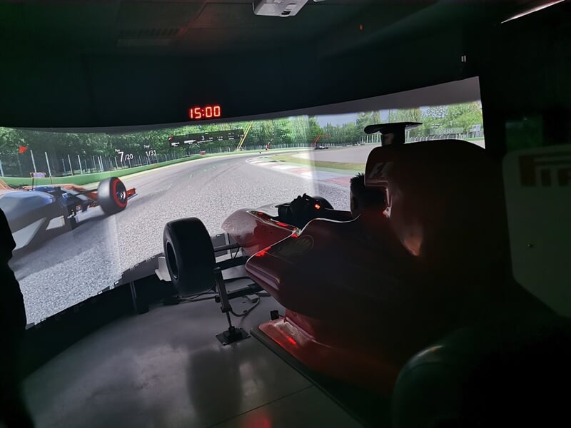 Simulateur de conduite automobile avec deux voitures rouges sur circuit virtuel, ambiance sombre et écran panoramique.