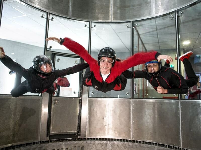 Trois personnes flottant dans un tunnel aérodynamique, participant central souriant en combinaison rouge.