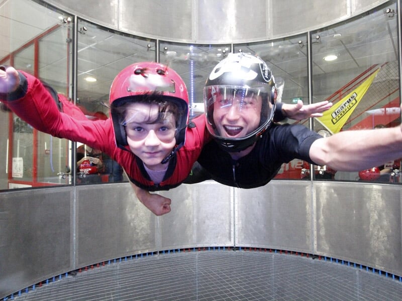 Enfant en combinaison rouge volant dans une soufflerie, assistée par un instructeur souriant.